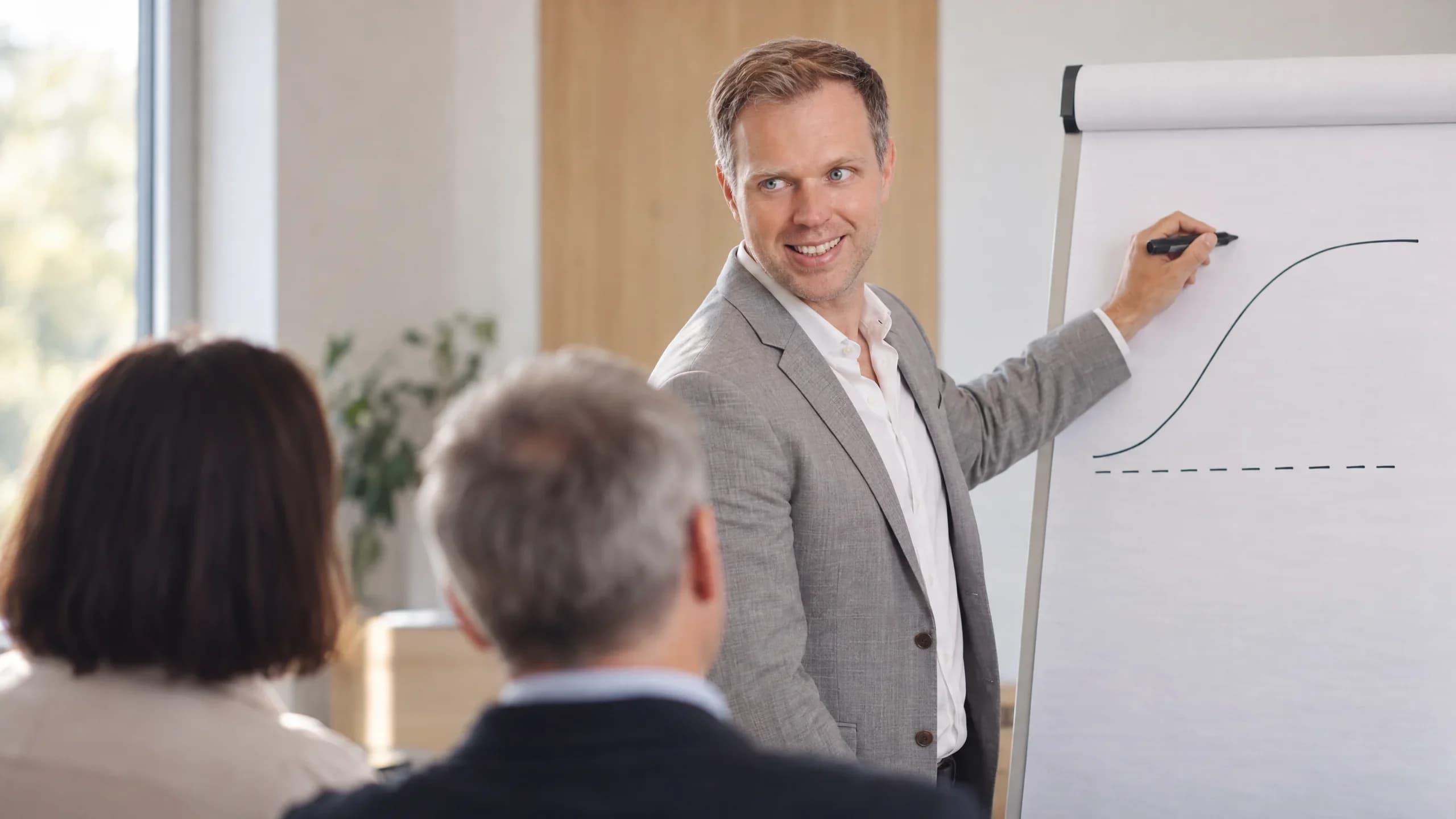 Dennis Tefett zeichnet am Flipchart eine ansteigende Kurve und wendet sich dabei lächelnd zu zwei Zuhörenden um