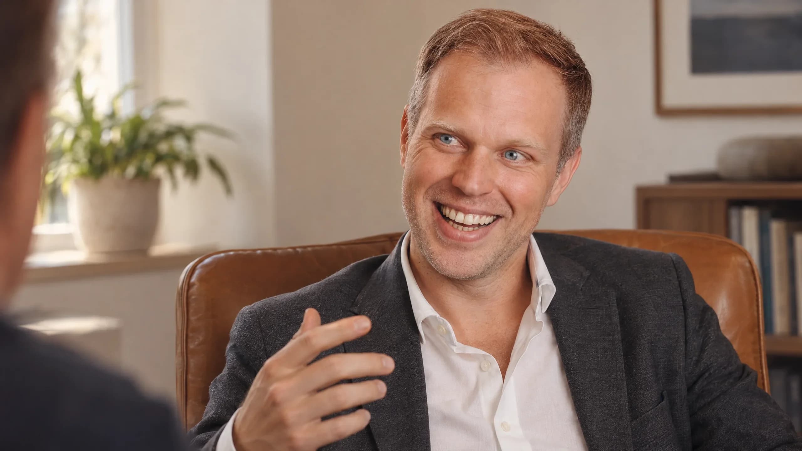 Dennis Tefett lacht im Ledersessel herzlich, eine Hand erklärend erhoben, sein Gegenüber ist nur am Bildrand zu sehen