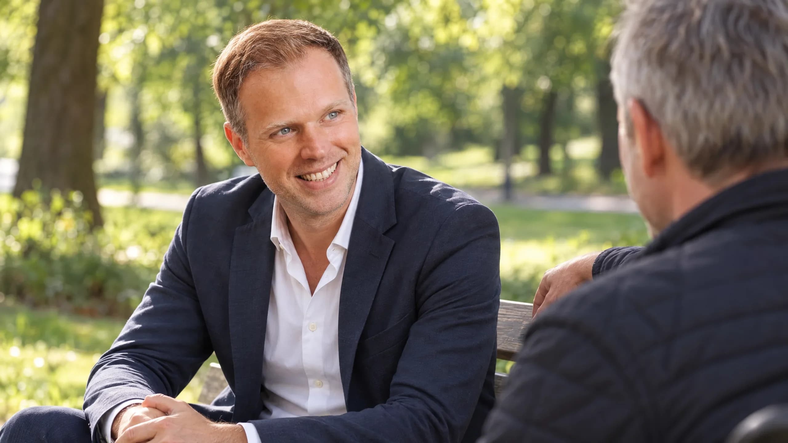Dennis Tefett sitzt auf einer Parkbank im sommerlichen Grün und lacht seinem Gesprächspartner zu, der ihm den Rücken zuwendet